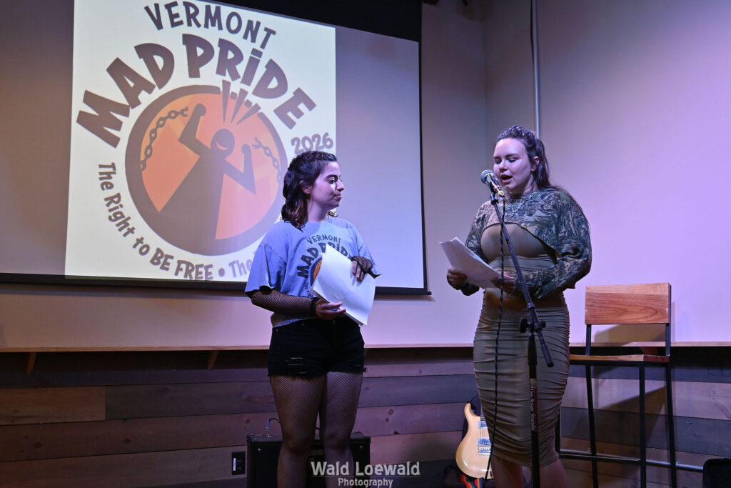 Neilah and Esme, MadFreedom Advocates staff, standing on stage with mic in front of Mad Pride 2026 logo