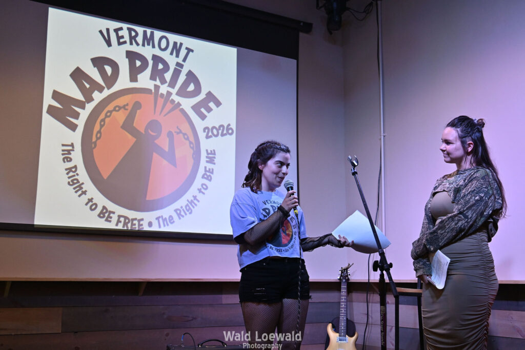 Neilah and Esme, MadFreedom Advocates staff, standing on stage with mic in front of Mad Pride 2026 logo