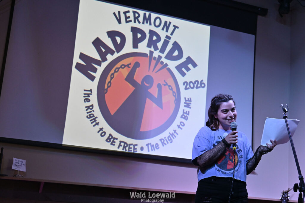 Neilah, MadFreedom Advocates staff, standing on stage with mic in front of Mad Pride 2026 logo
