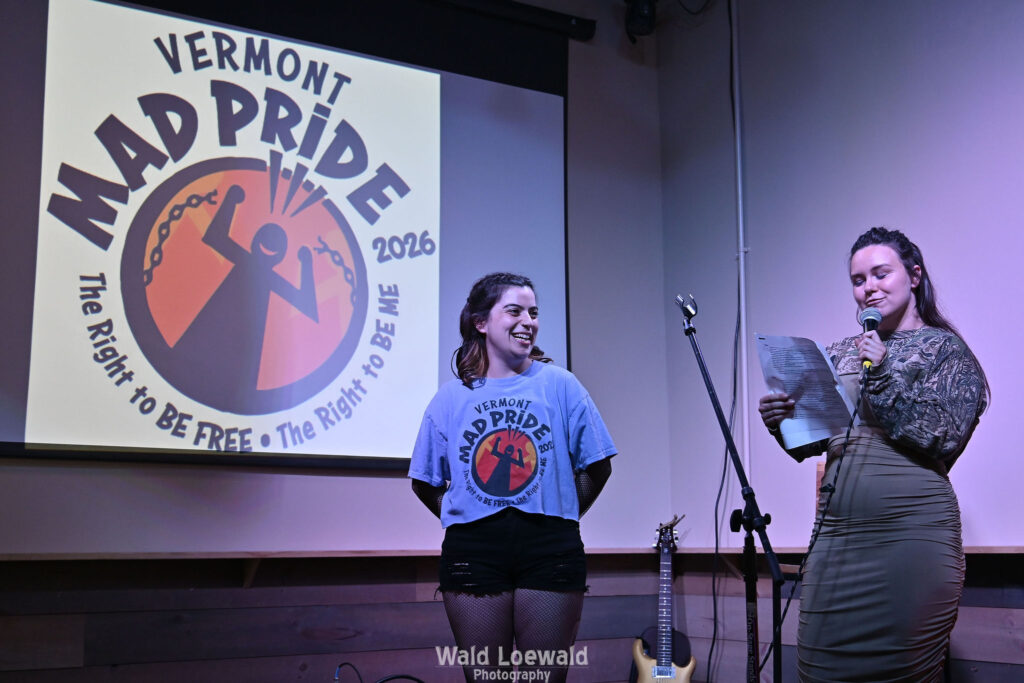 Neilah and Esme, MadFreedom Advocates staff, standing on stage with mic in front of Mad Pride 2026 logo