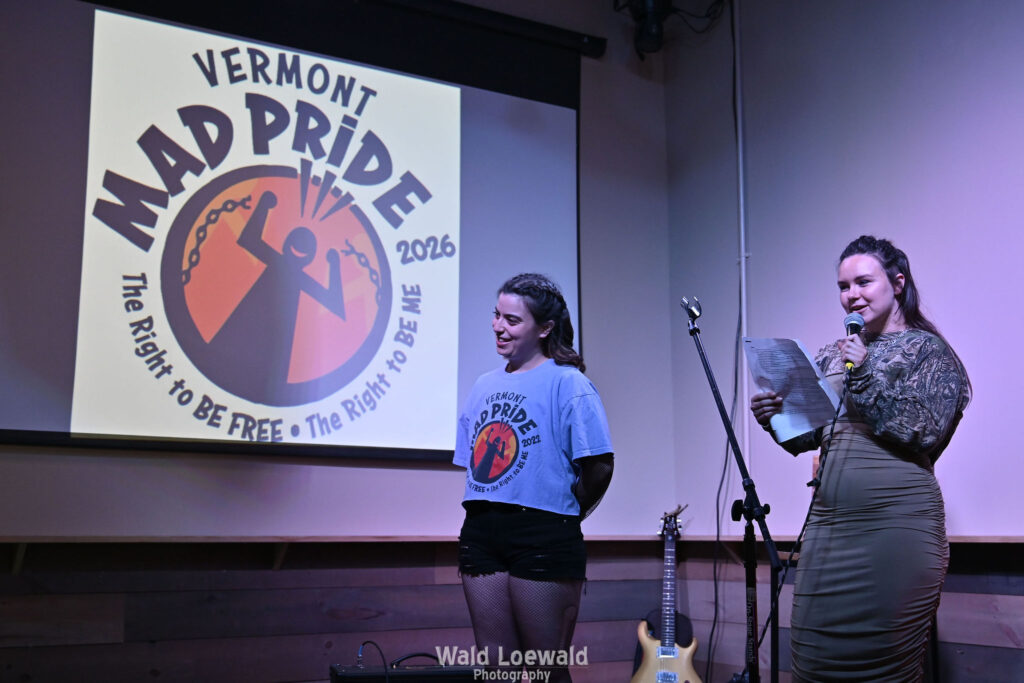 Neilah and Esme, MadFreedom Advocates staff, standing on stage with mic in front of Mad Pride 2026 logo