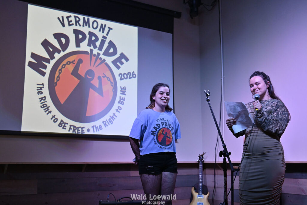 Neilah and Esme, MadFreedom Advocates staff, standing on stage with mic in front of Mad Pride 2026 logo