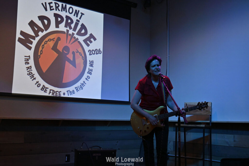 Performer playing guitar behind a mic in front of the Mad Pride 2026 logo