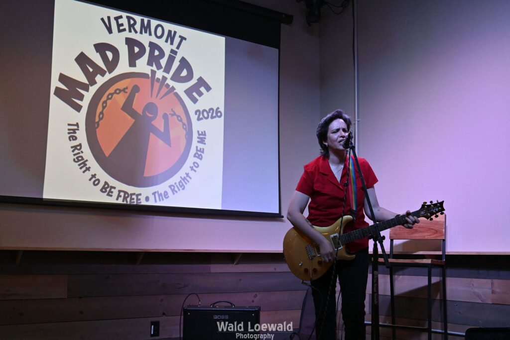 Performer playing guitar behind a mic in front of the Mad Pride 2026 logo