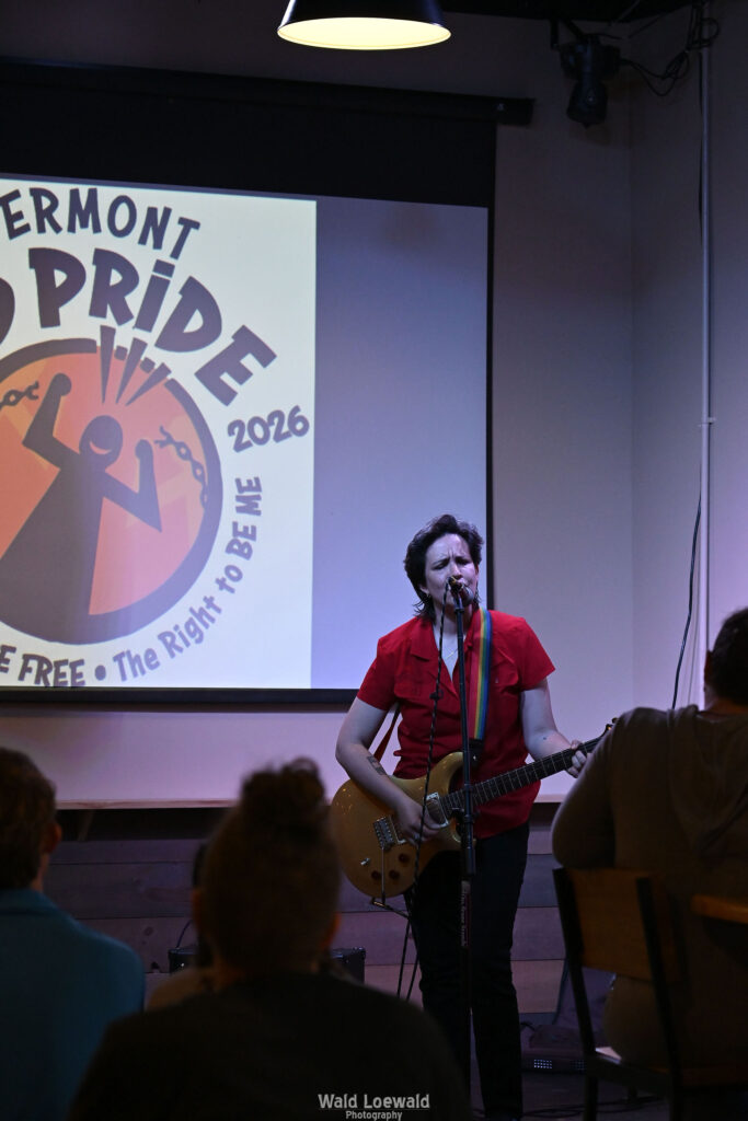 Performer playing guitar behind a mic in front of the Mad Pride 2026 logo