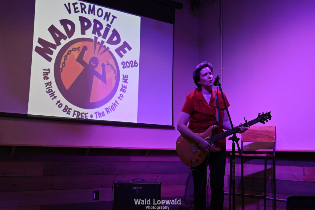 Performer playing guitar behind a mic in front of the Mad Pride 2026 logo