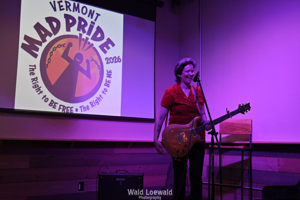 Performer playing guitar behind a mic in front of the Mad Pride 2026 logo