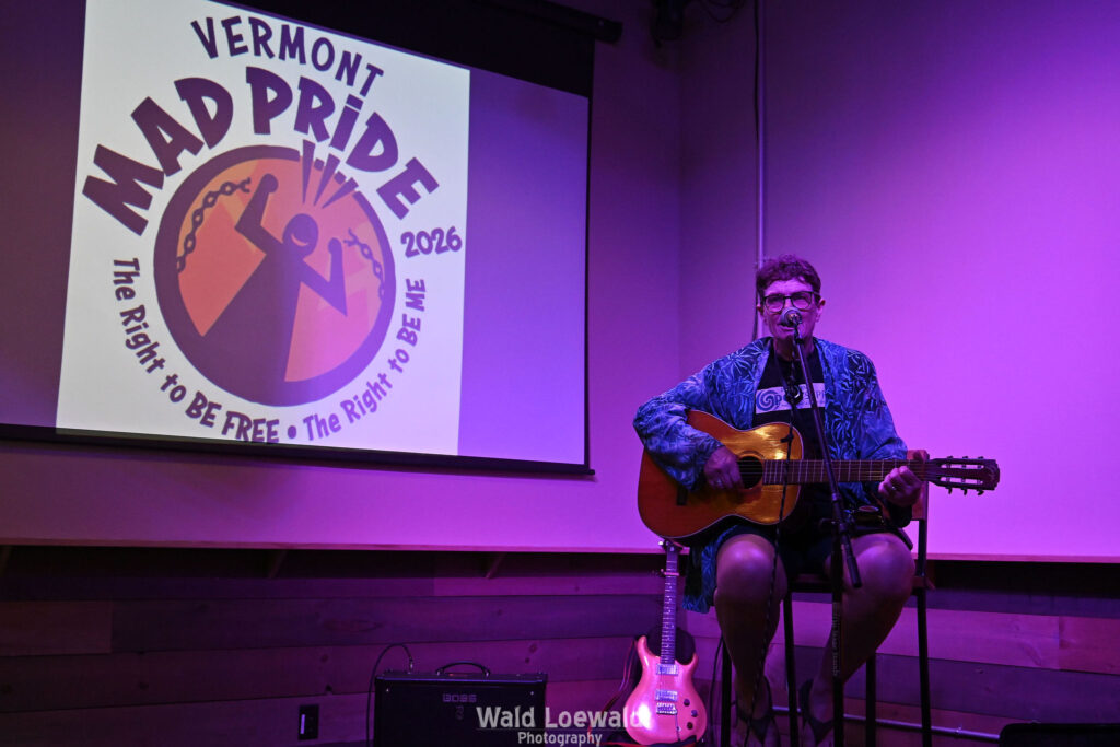 Chris Hansen sitting behind a mic playing guitar, in front of a Mad Pride 2026 logo