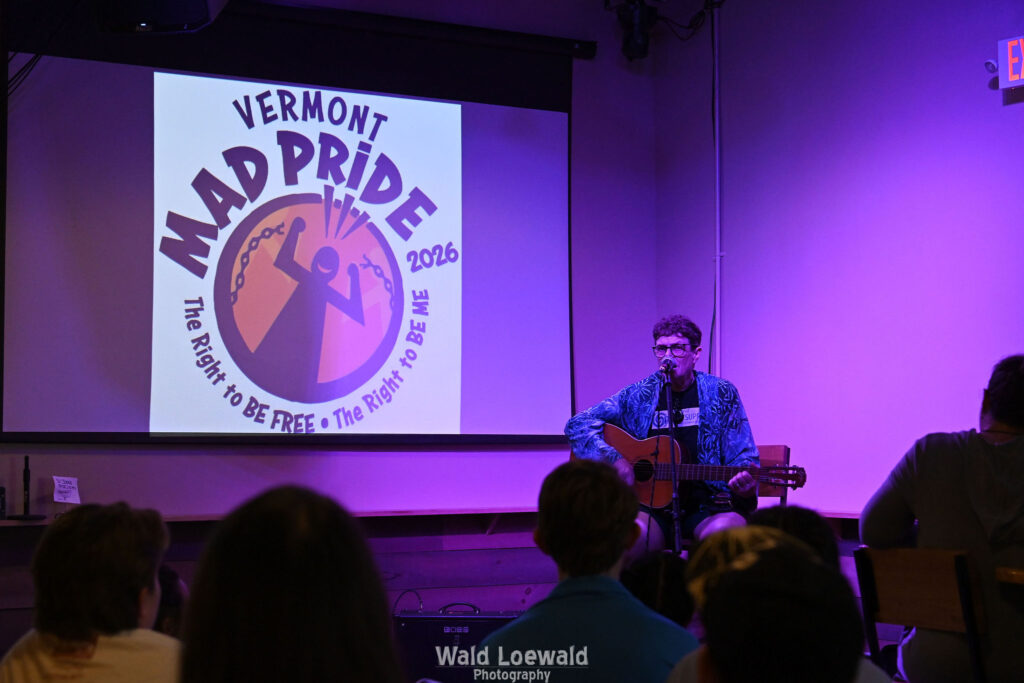 Chris Hansen sitting behind a mic playing guitar, in front of a Mad Pride 2026 logo