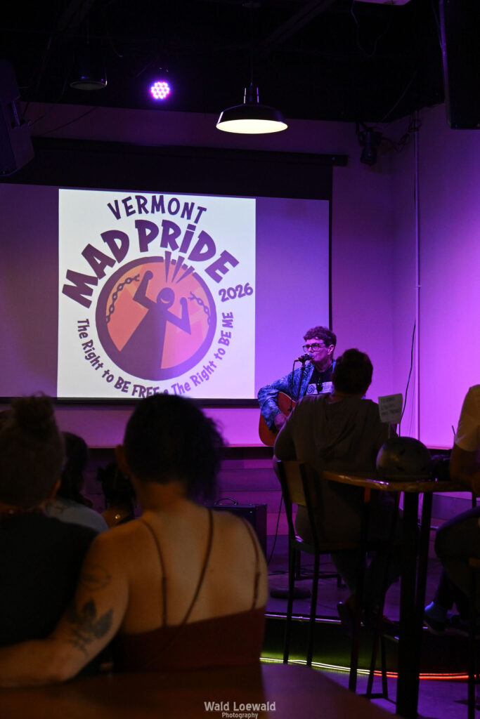 Chris Hansen sitting behind a mic playing guitar, in front of a Mad Pride 2026 logo
