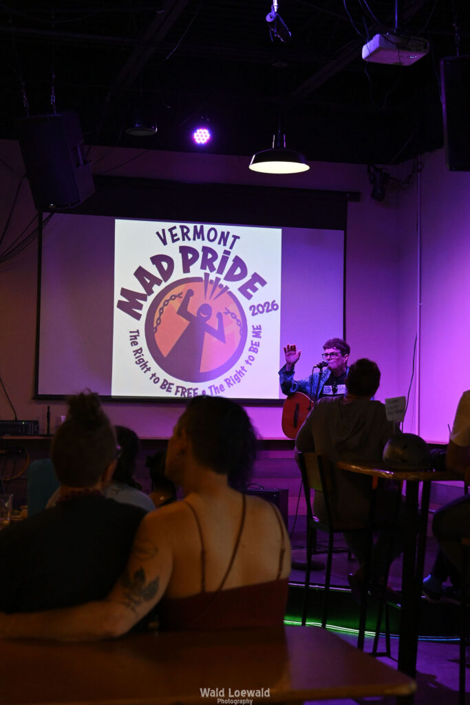 Chris Hansen sitting behind a mic playing guitar, in front of a Mad Pride 2026 logo