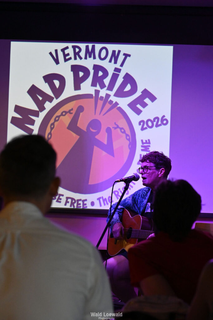 Chris Hansen sitting behind a mic playing guitar, in front of a Mad Pride 2026 logo