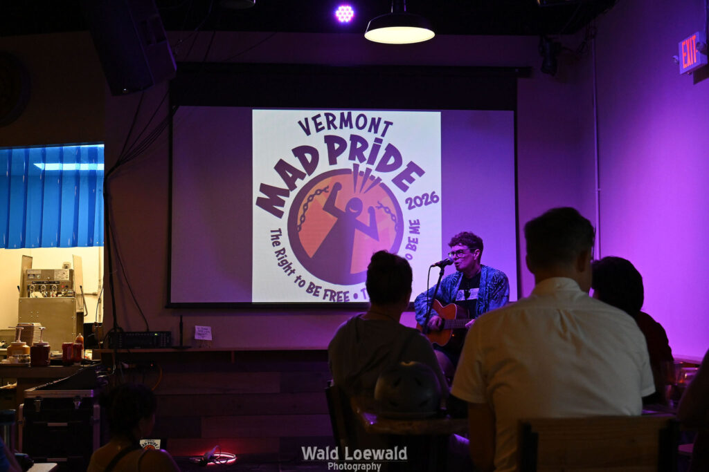 Chris Hansen sitting behind a mic playing guitar, in front of a Mad Pride 2026 logo