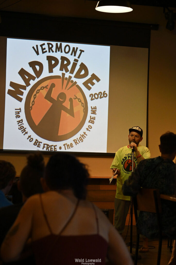 Cal Moen standing behind a mic and in front of a Mad Pride 2026 Logo