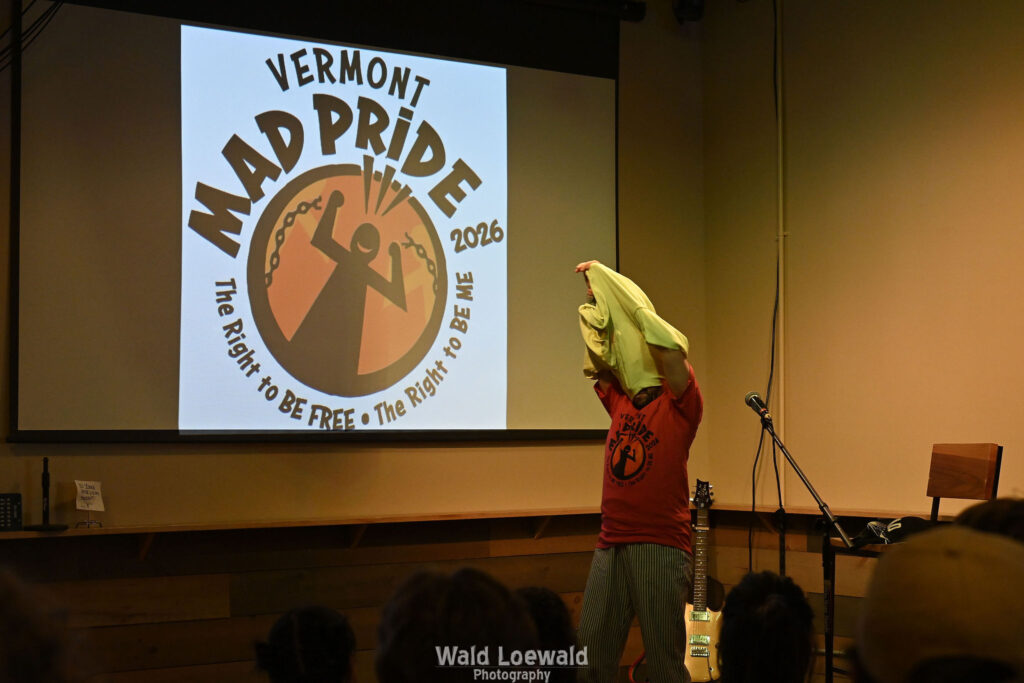 Cal Moen standing behind a mic and in front of a Mad Pride 2026 Logo