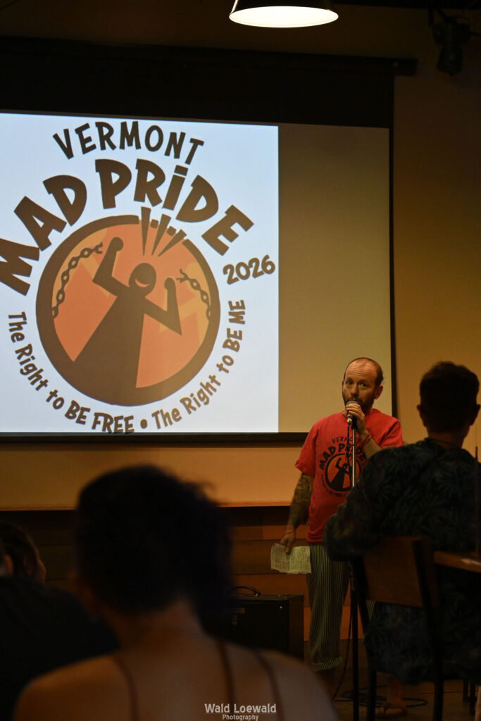 Cal Moen standing behind a mic and in front of a Mad Pride 2026 Logo