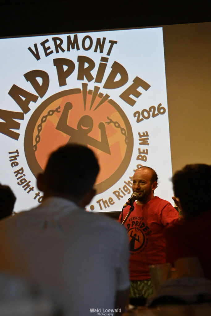 Cal Moen standing behind a mic and in front of a Mad Pride 2026 Logo