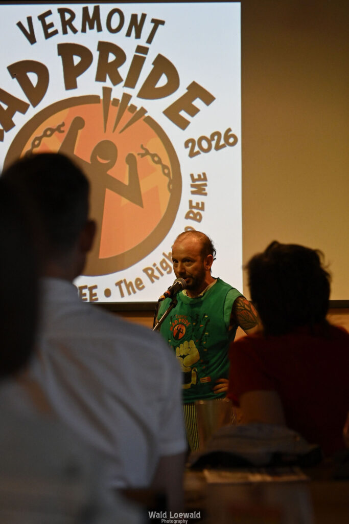 Cal Moen standing behind a mic and in front of a Mad Pride 2026 Logo