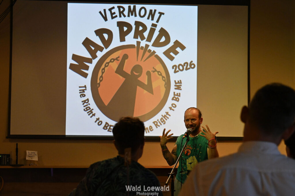 Cal Moen standing behind a mic and in front of a Mad Pride 2026 Logo