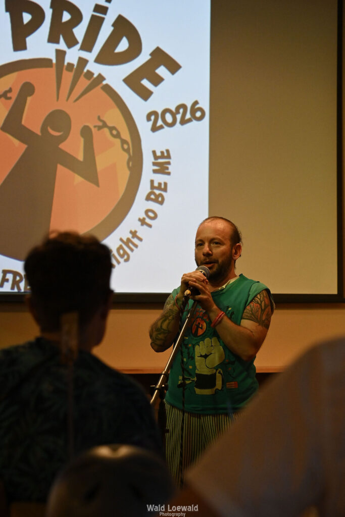Calvin Moen in old Mad pride shirt in front of mic with yellow lighting and Mad Pride 2026 logo