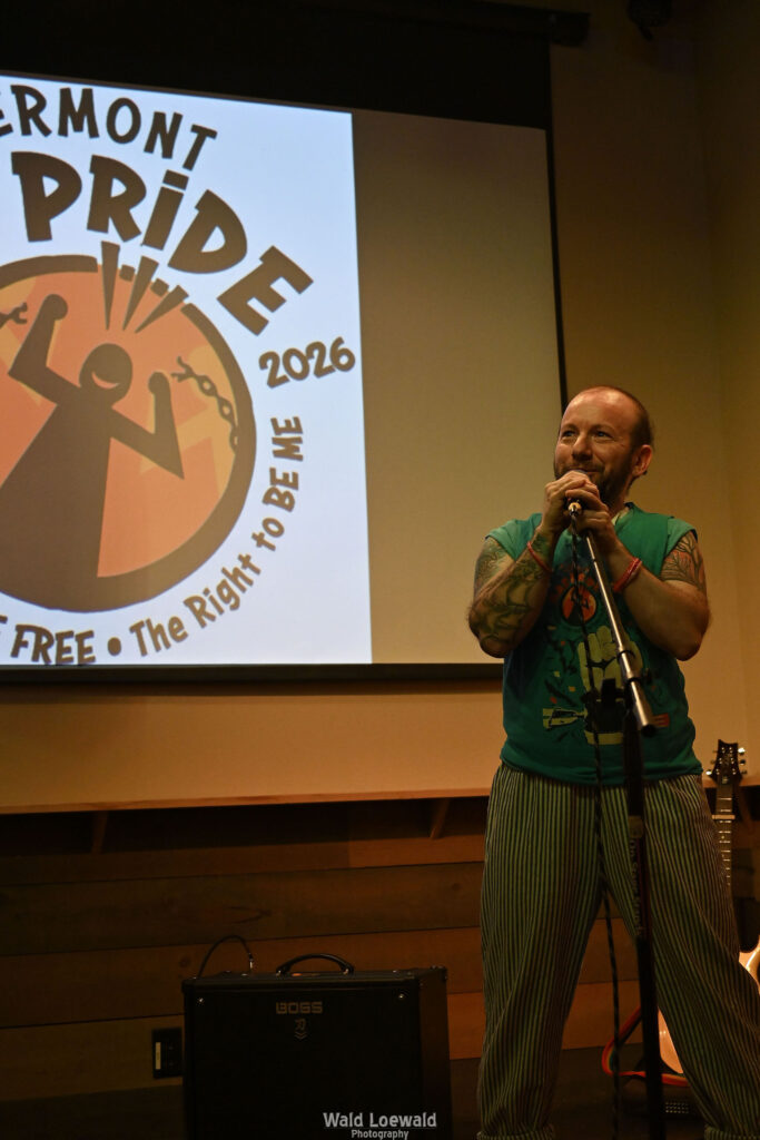 Calvin Moen in old Mad pride shirt in front of mic with yellow lighting and Mad Pride 2026 logo