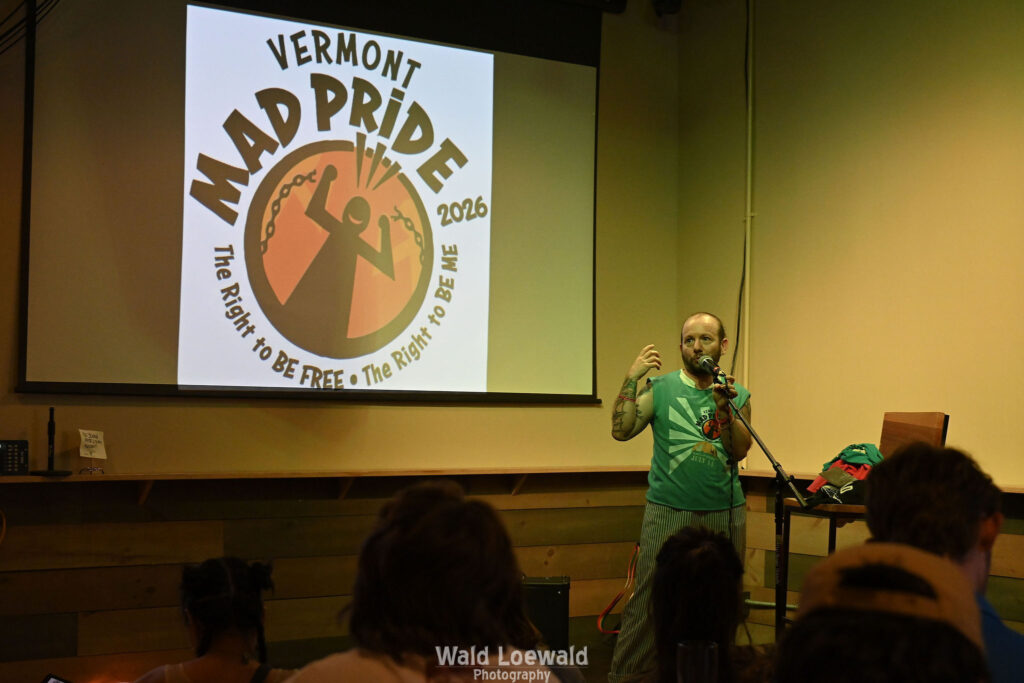 Calvin Moen in old Mad pride shirt in front of mic with yellow lighting and Mad Pride 2026 logo