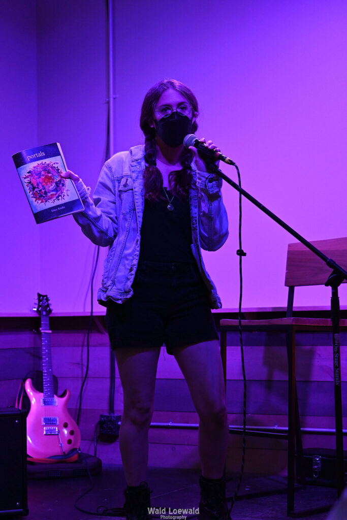 Performer on stage with mic in pink lighting with a book and a mask