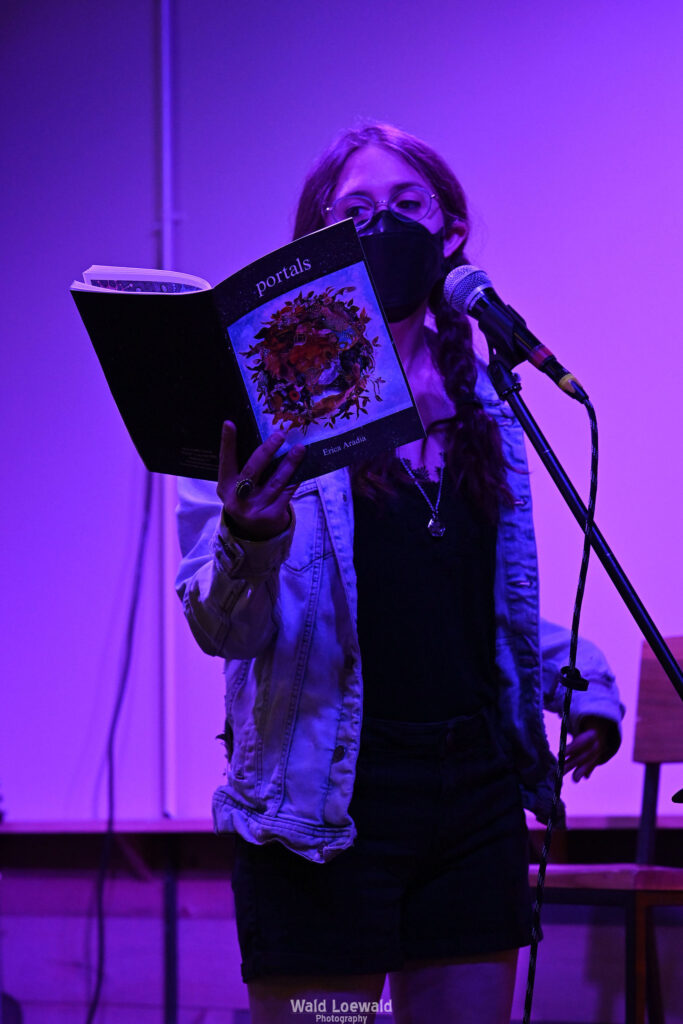 Performer on stage with mic in pink lighting with a book and a mask