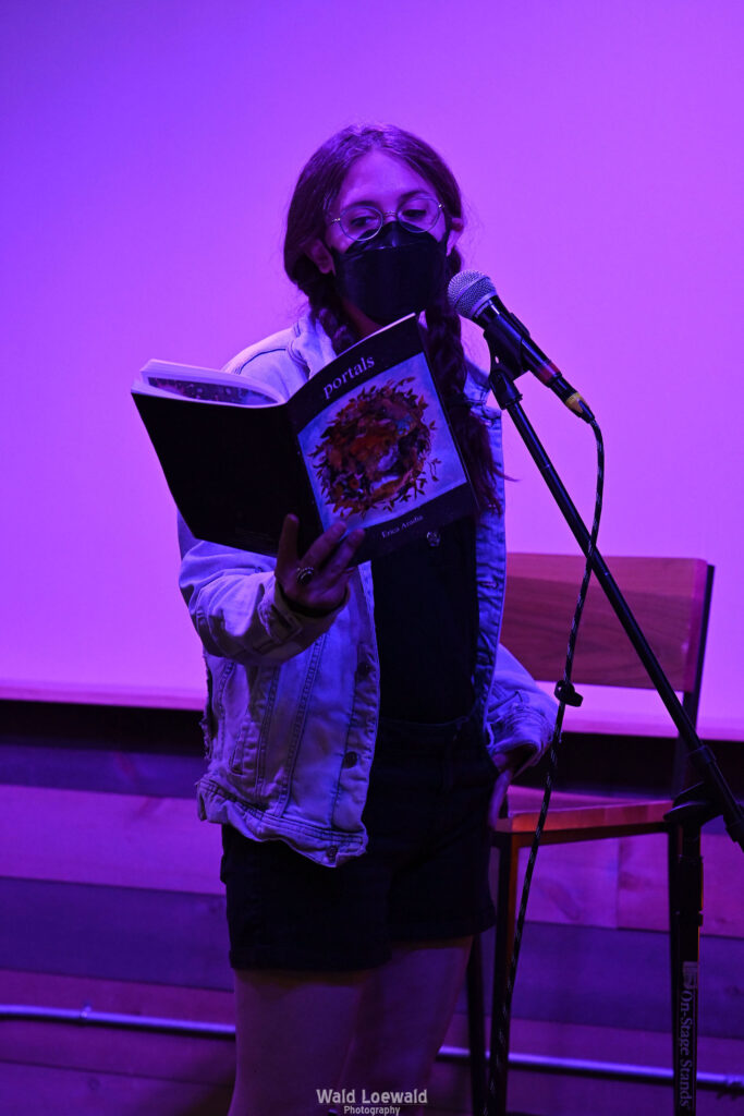 Performer on stage with mic in pink lighting with a book and a mask in front of Mad Pride 2026 logo