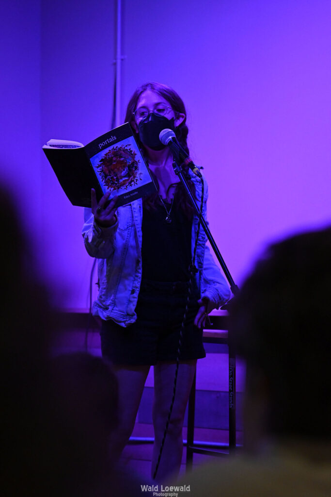 Performer on stage with mic in pink lighting with a book and a mask in front of Mad Pride 2026 logo