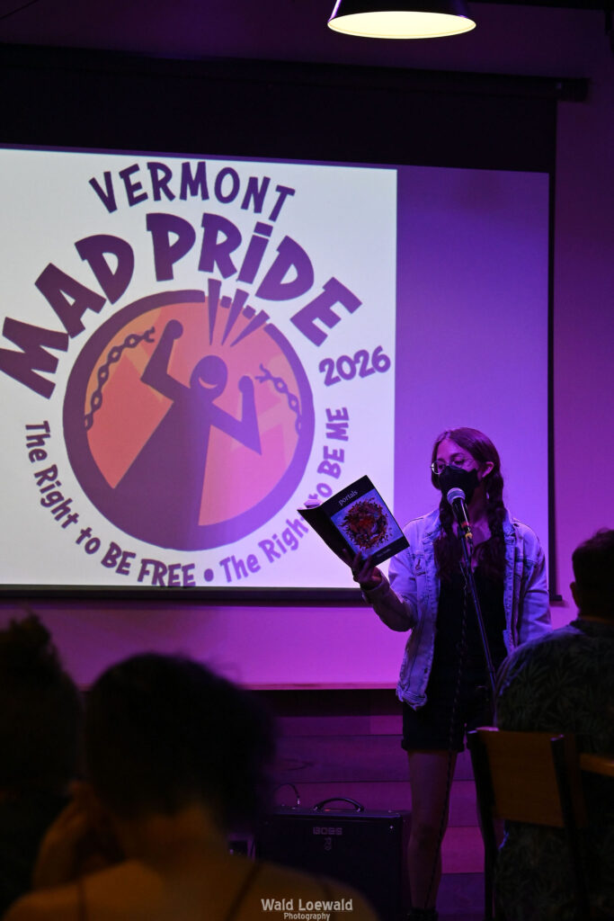 Performer on stage with mic in pink lighting with a book and a mask in front of Mad Pride 2026 logo