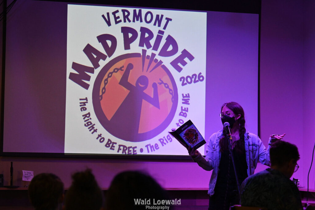 Performer on stage with mic in pink lighting with a book and a mask in front of Mad Pride 2026 logo