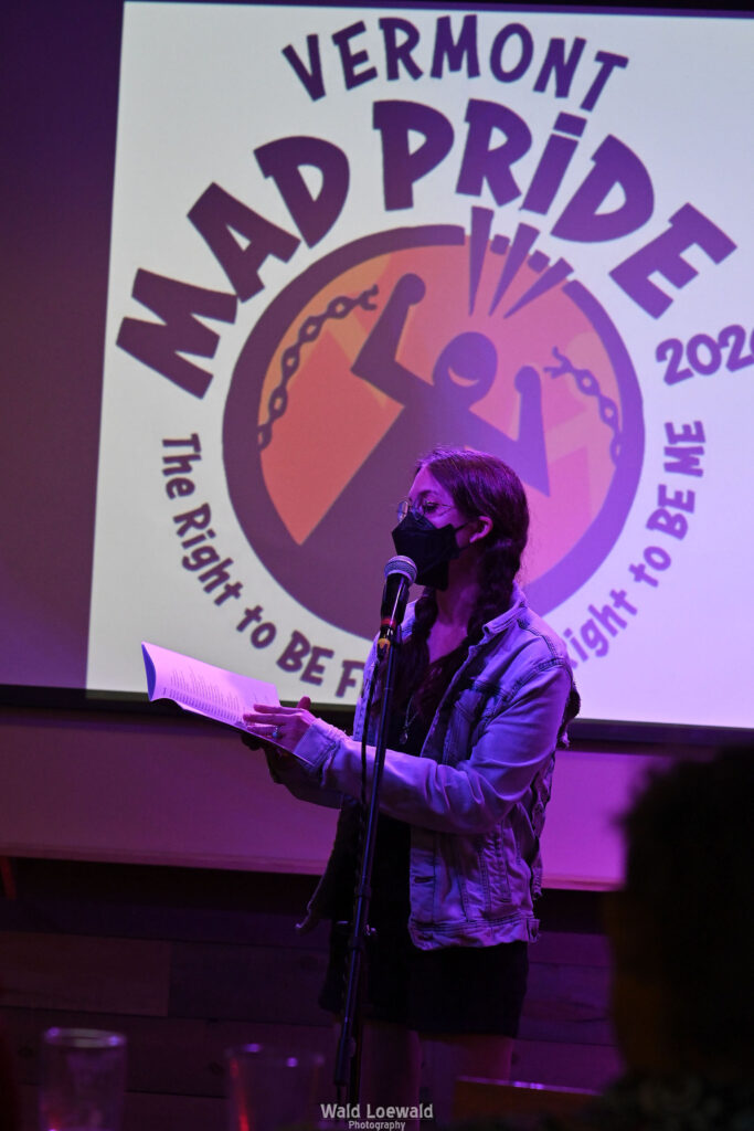 Performer on stage with mic in pink lighting with a book and a mask in front of Mad Pride 2026 logo