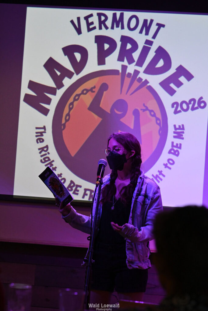 Performer on stage with mic in pink lighting with a book and a mask in front of Mad Pride 2026 logo
