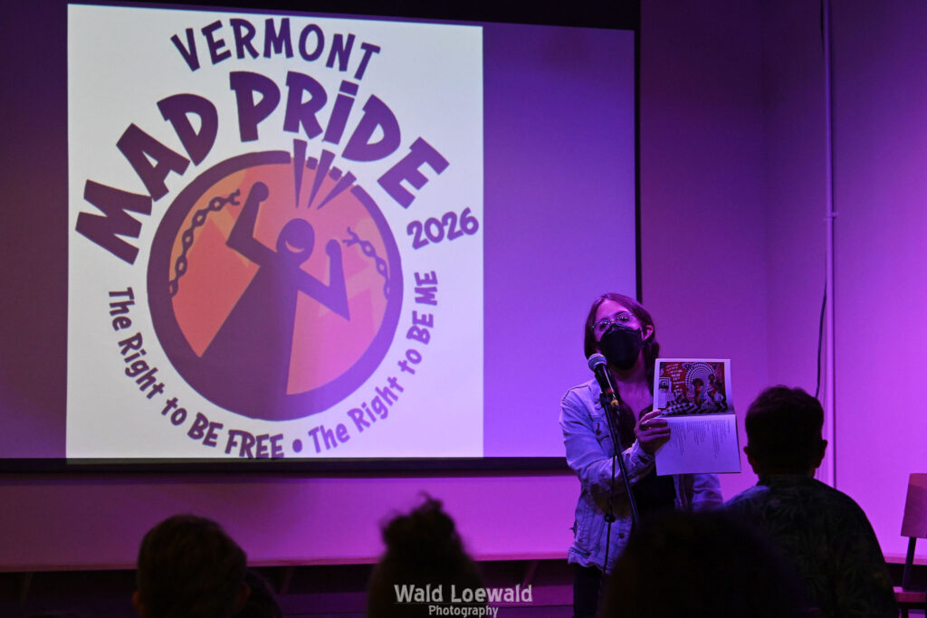 Performer on stage with mic in pink lighting with a book and a mask in front of Mad Pride 2026 logo