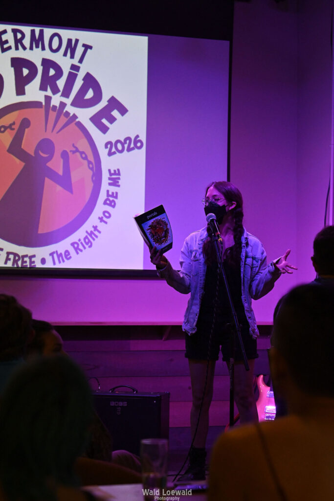 Performer on stage with mic in pink lighting with a book and a mask in front of Mad Pride 2026 logo