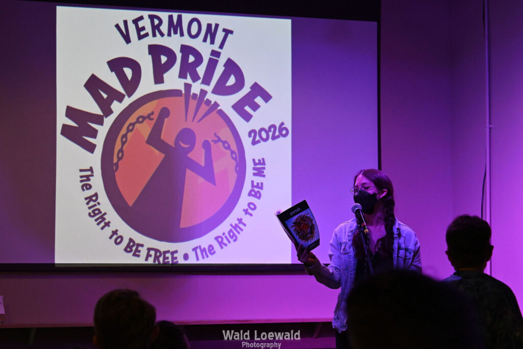Performer on stage with mic in pink lighting with a book and a mask in front of Mad Pride 2026 logo