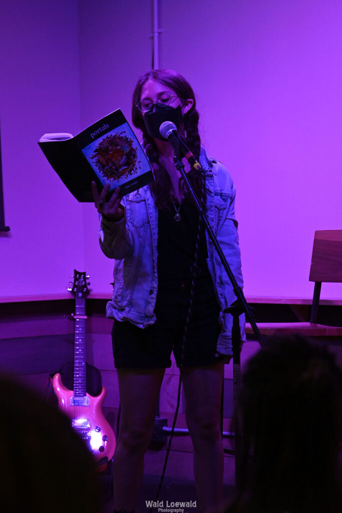 Performer on stage with mic in pink lighting with a book and a mask