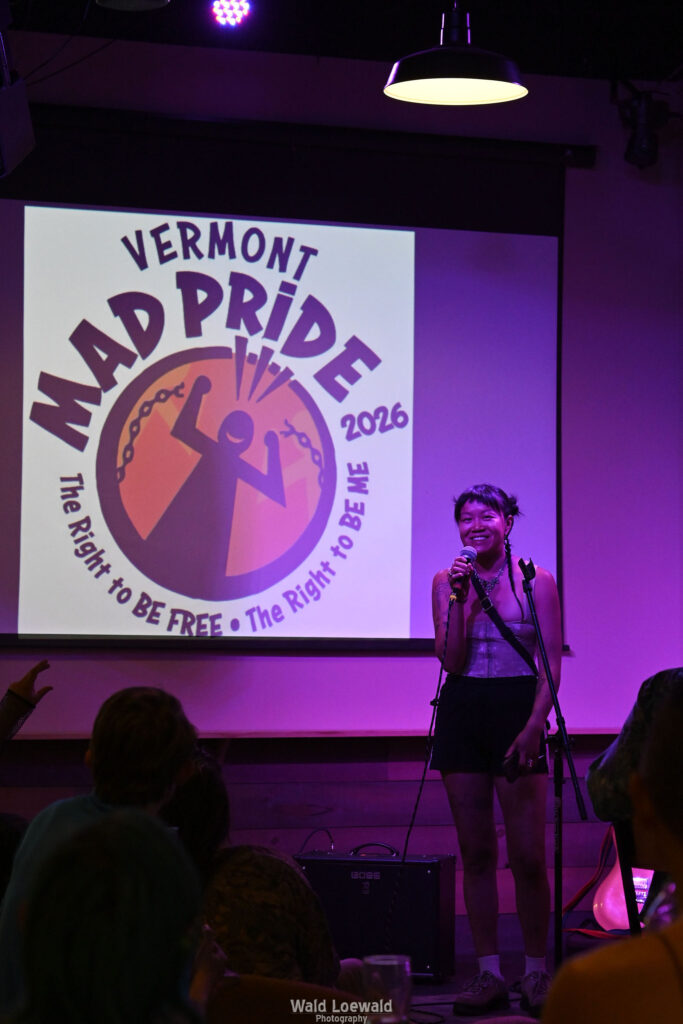 Performer on stage with mic in pink lighting in front of Mad Pride 2026 logo