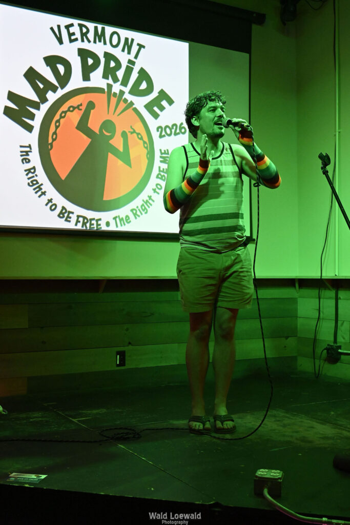 Chris Nial in green lighting holding a mic in front of Mad Pride 2026 logo