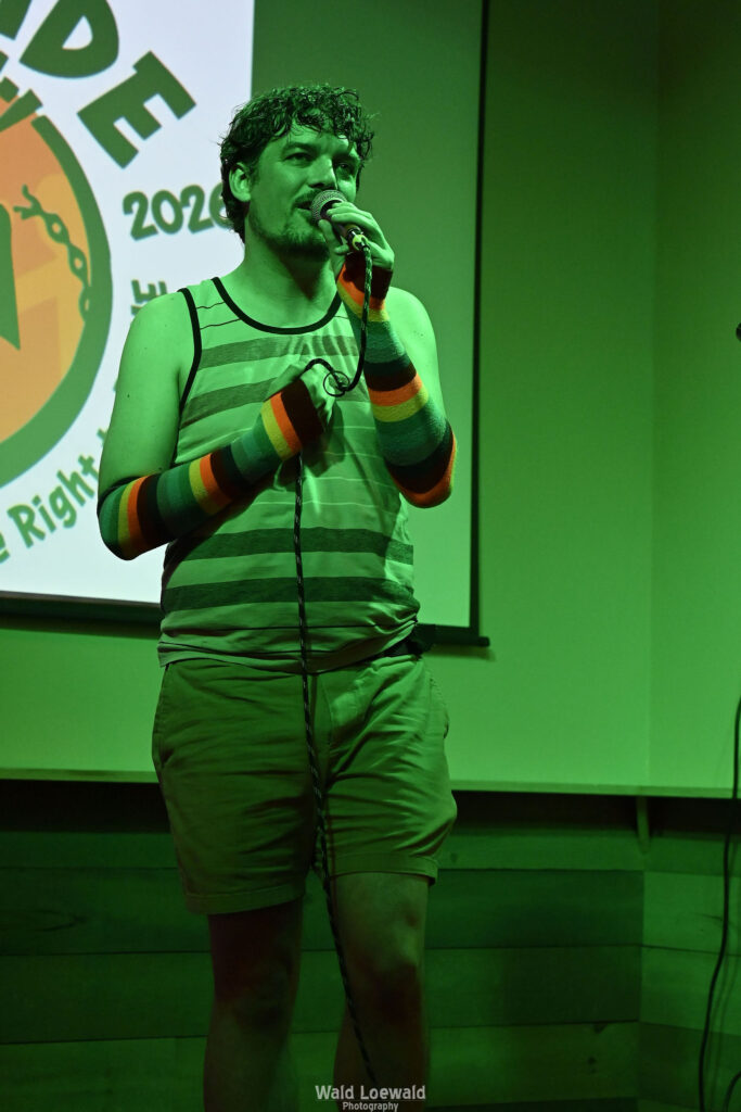 Chris Nial in green lighting holding a mic in front of Mad Pride 2026 logo