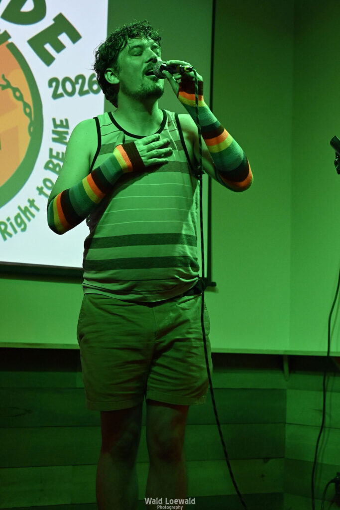 Chris Nial in green lighting holding a mic in front of Mad Pride 2026 logo