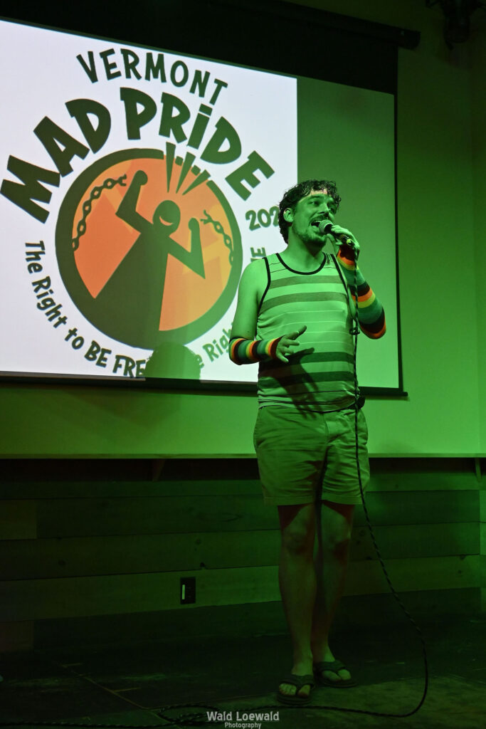Chris Nial in green lighting holding a mic in front of Mad Pride 2026 logo