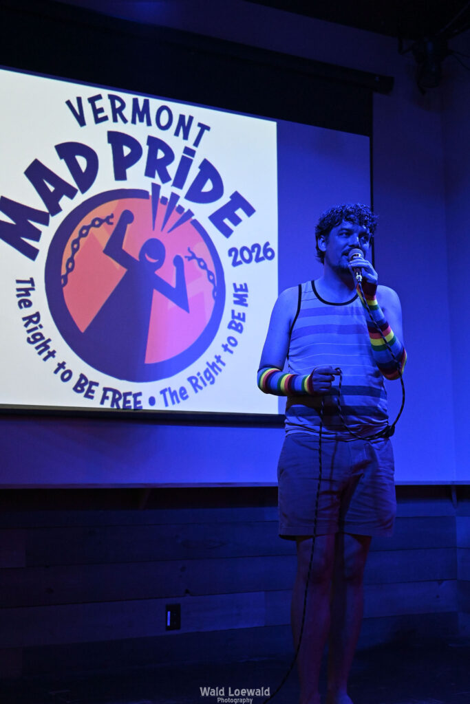 Chris Nial in blue lighting holding a mic in front of Mad Pride 2026 logo