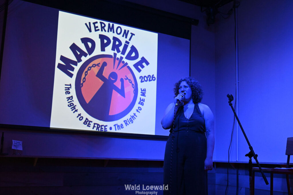Performer in blue lighting holding a mic in front of Mad Pride 2026 logo