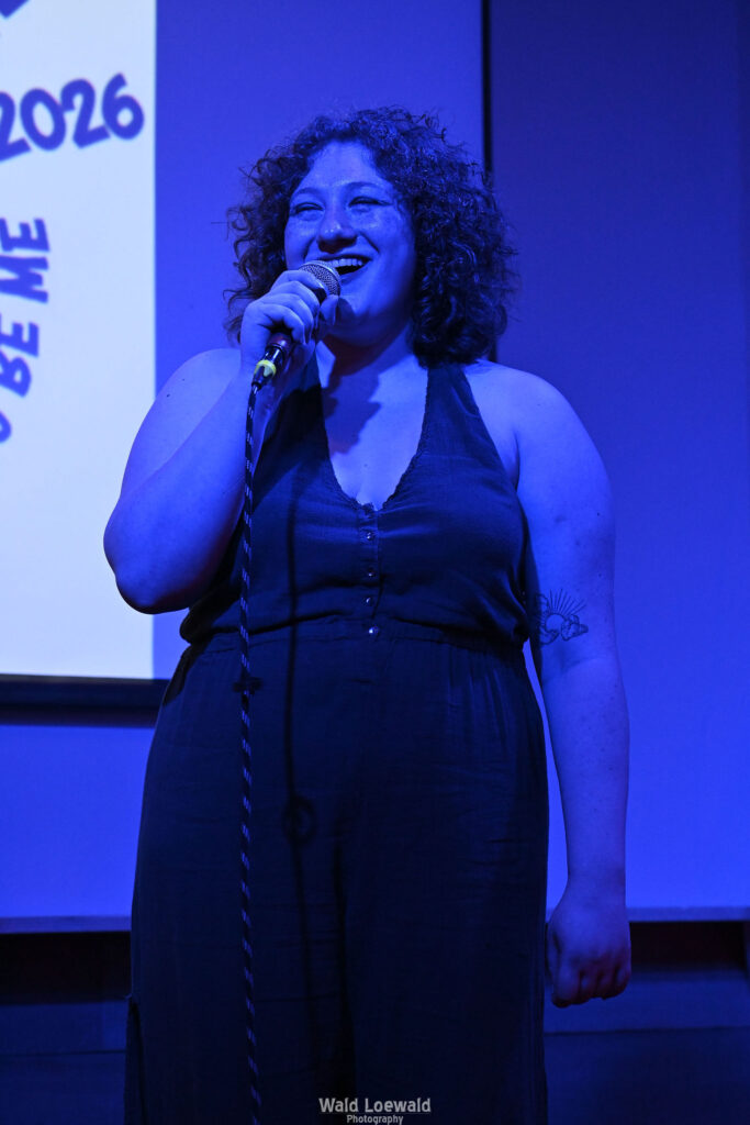 Performer in blue lighting holding a mic in front of Mad Pride 2026 logo, smiling