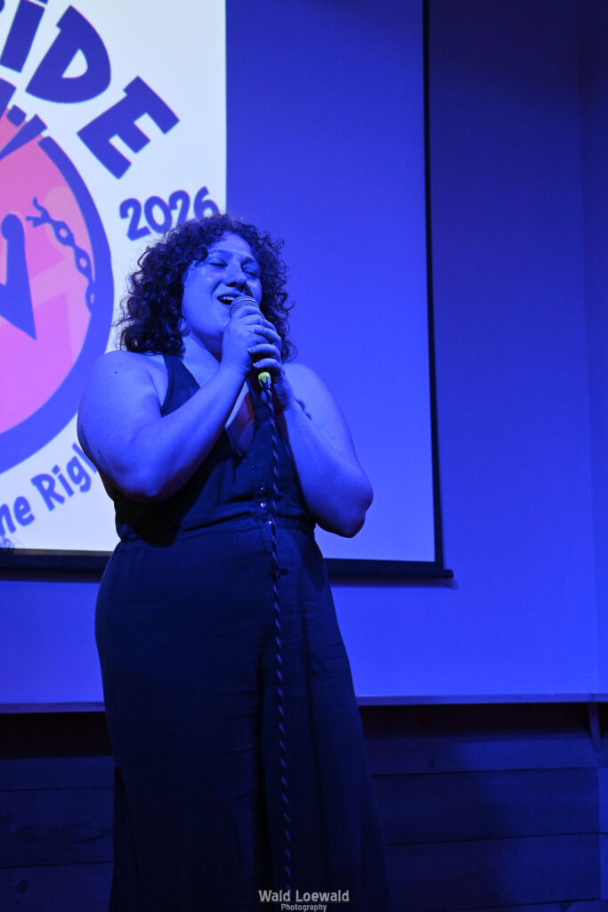 Performer in blue lighting holding a mic in front of Mad Pride 2026 logo