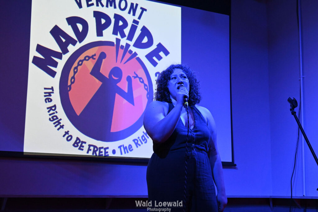 Performer in blue lighting holding a mic in front of Mad Pride 2026 logo