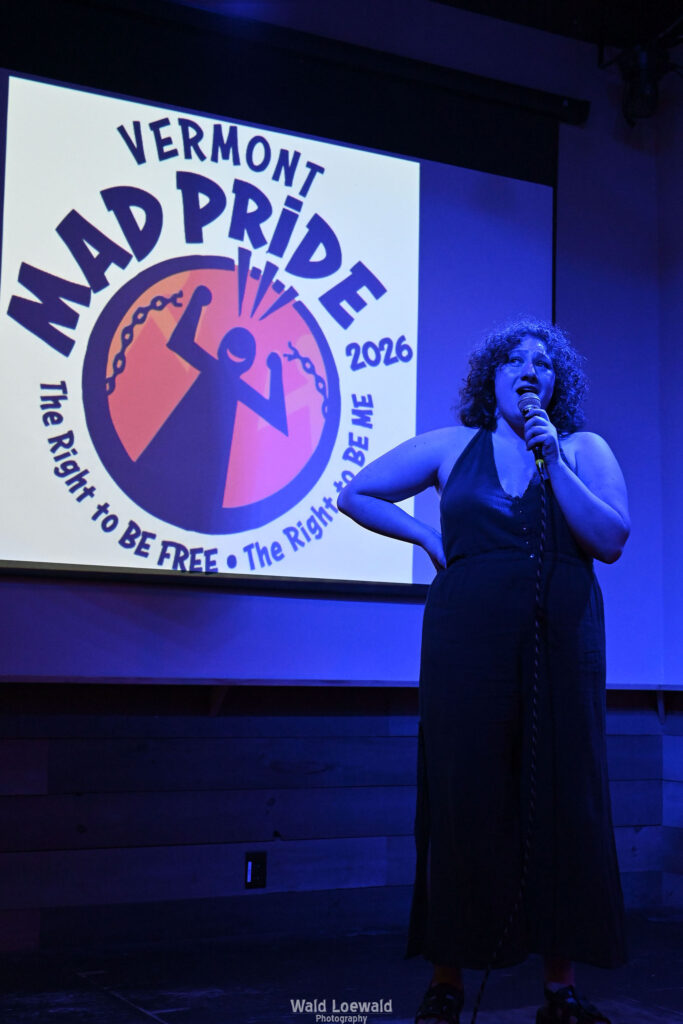 Performer in blue lighting holding a mic in front of Mad Pride 2026 logo