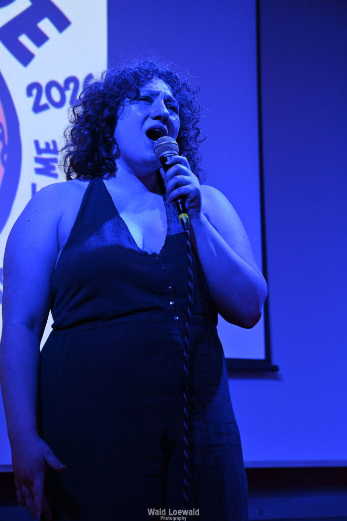 Performer in blue lighting holding a mic in front of Mad Pride 2026 logo